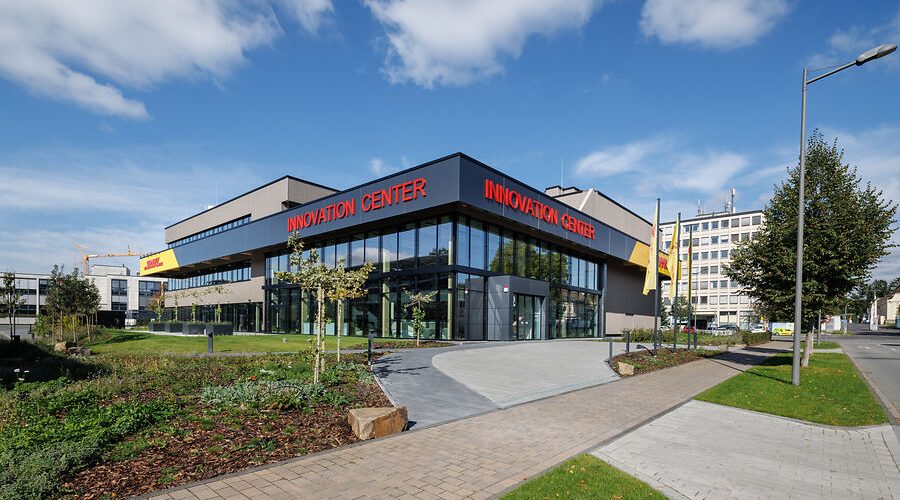 Modernes Gebäude mit der Aufschrift Innovation Center in roten Buchstaben, große Glasfassade, umgeben von Grünflächen und Wegen, bei sonnigem Wetter und blauem Himmel.